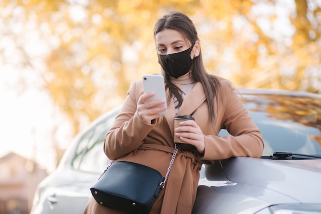 Attraktive junge Frau in Schutzmaske mit Tasse Kaffee stehen neben dem Auto und benutzen Telefon.
