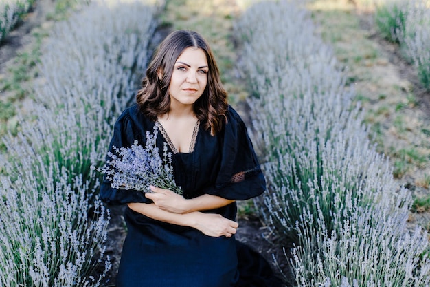 Attraktive junge Frau in schwarzem Kleid, die von einem Lavendelfeld umgeben ist und einen Blumenstrauß hält