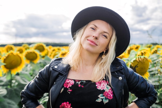 Attraktive junge Frau in schwarzer Lederjacke und Hut posiert auf einem Sonnenblumenfeld