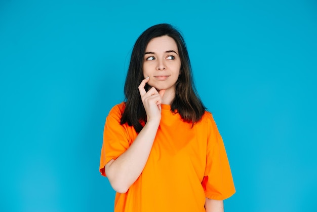 Attraktive junge Frau in trendiger orangefarbener Kleidung, nachdenklicher und skeptischer Gesichtsausdruck, leerer Raum für Kopien, isoliert auf blauem Hintergrund, Porträtfoto