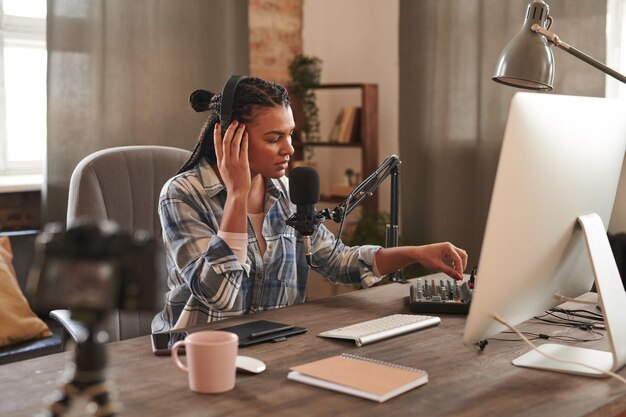 Foto attraktive junge frau mit afro-zöpfen sitzt am schreibtisch im loft-heimstudio und passt die mikrofoneinstellungen an, um mit den videoaufnahmen für einen blog zu beginnen