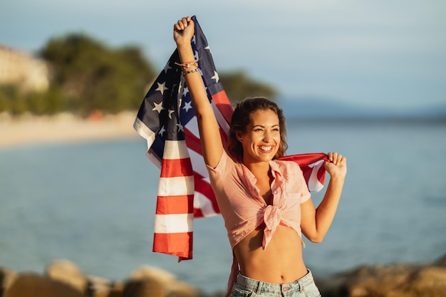 Attraktive junge frau mit amerikanischer nationalflagge genießt einen entspannten tag am strand.