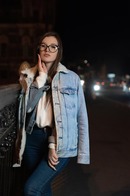 Attraktive junge Frau mit Brille und Jeansjacke auf abendlichem Stadthintergrund Stilvolles Mädchen auf nächtlicher Stadtstraße