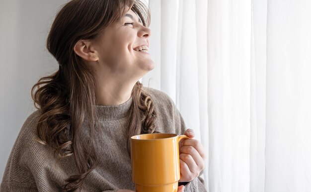 Attraktive junge Frau mit einer Tasse heißem Getränk am Fenster