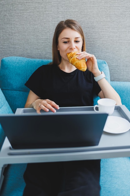 Attraktive junge Frau mit Laptop-Computer, während sie zu Hause auf dem Sofa mit Kaffee und Croissants sitzt Arbeiten Sie online aus der Ferne