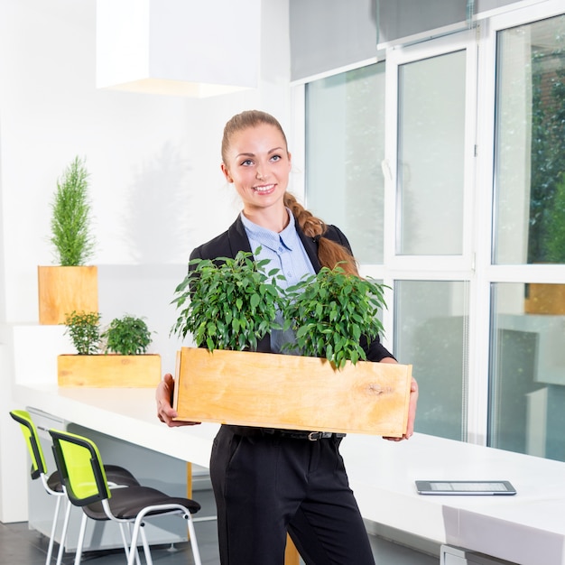 Attraktive junge Geschäftsfrau, die im modernen Dachbodenbüro steht, das eine Box mit Pflanzen hält