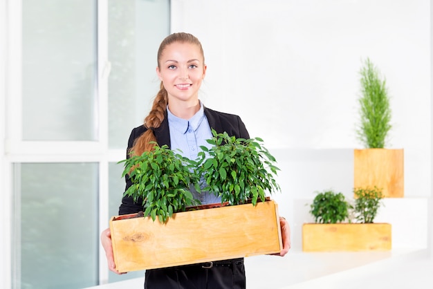 Attraktive junge Geschäftsfrau, die im modernen Dachbodenbüro steht, das eine Box mit Pflanzen hält