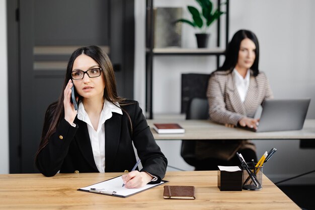Attraktive junge Geschäftsfrau mit Jacke, die am Handy spricht, während sie auf einem Schreibtisch sitzt und ein Notizbuch in der Nähe ihres Kollegen bei der Arbeit im Büro benutzt