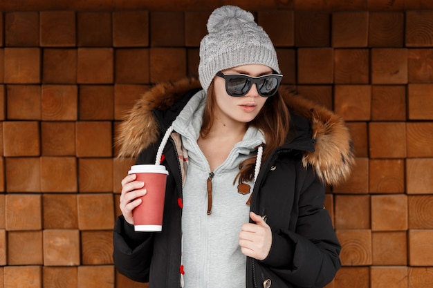 Foto attraktive junge hipster-frau in strickmütze in einem sweatshirt in einer stilvollen jacke in sonnenbrille, die nahe einer hölzernen weinlesewand aufwirft und eine tasse kaffee in der hand hält. netter mädchenwintertag