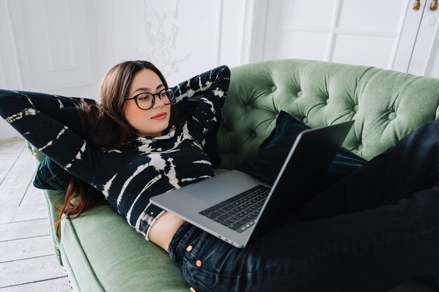 Attraktive junge kaukasische Frau, die auf einem Sofa ruht und Laptop-Computer verwendet.