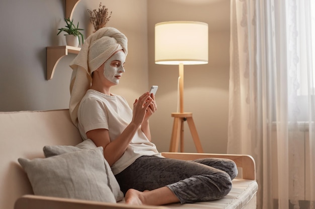Attraktive junge kaukasische Frau, die mit ihrem Handy in ein Handtuch und eine Tonmaske im Gesicht gewickelt ist, während sie sich auf dem Sofa in ihrem Wohnzimmer entspannt und kosmetische Eingriffe durchführt