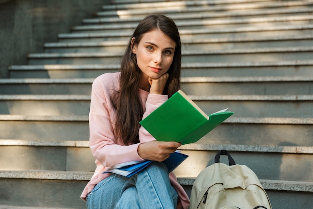 Attraktive junge lächelnde Studentin, die ein Buch liest, während sie auf der Treppe sitzt