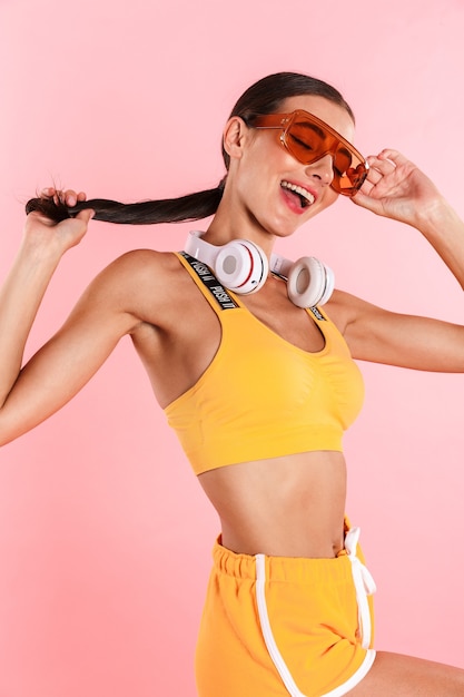 Attraktive junge schlanke brünette Frau in Sportkleidung, Kopfhörer tragend, isoliert über rosa Wand posieren