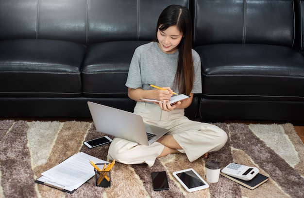 Attraktive junge schöne asiatische Frau Unternehmerin oder Freiberuflerin, die zu Hause mit Laptop-Geschäftsberichten und Online-Kommunikation auf Wohnzimmer-Sofa arbeitet und Fernzugriffskonzept arbeitet.