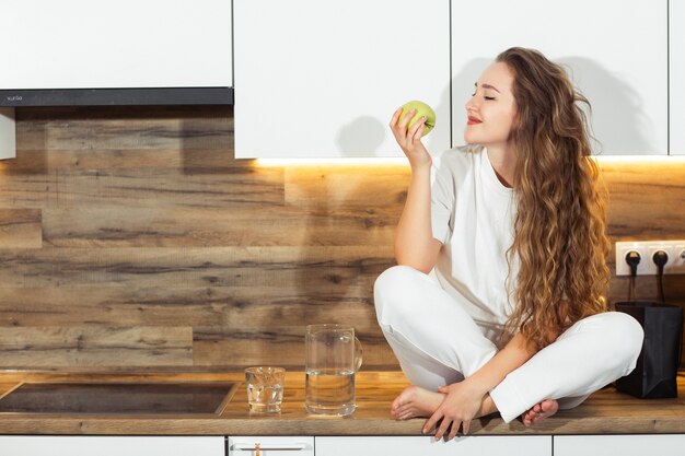 Attraktive junge weiße Frau, die einen grünen Apfel für ihr Frühstück am Morgen beißt. Gesundes Esskonzept .Diet. Schöne junge Frau, die auf Küchentisch sitzt. Früchte.