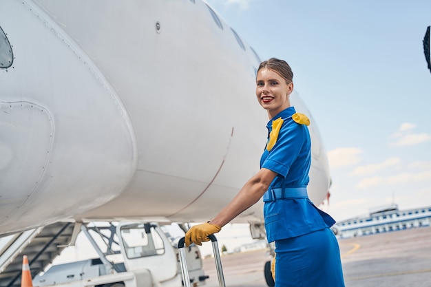 Attraktive kaukasische Dame in einer Flugbegleiteruniform, die ihre behandschuhte Hand auf den Gepäckgriff stützt