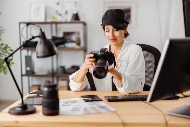 Attraktive kaukasische Frau in stilvoller Kleidung, die am Schreibtisch sitzt und mit Computer und Kamera arbeitet Konzept von Menschen und kreativem Hobby