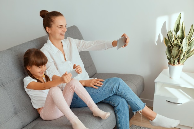 Attraktive kaukasische Frau mit Haarknoten, die weißes Hemd und Jeans trägt, die mit ihrer süßen kleinen Tochter auf dem Sofa sitzt und das Handy in der Hand hält, während sie während des Livestreams mit Anhängern spricht.