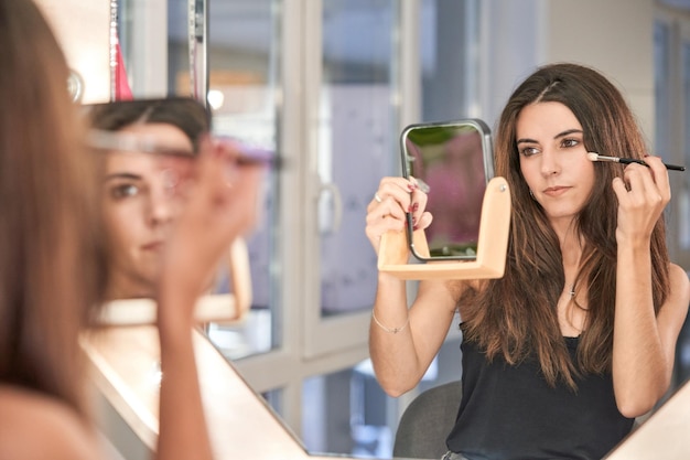 Attraktive Kundin mit langen Haaren blickt in den Spiegel, während sie während eines Termins im modernen, hellen Schönheitssalon Make-up mit einem Pinsel aufträgt