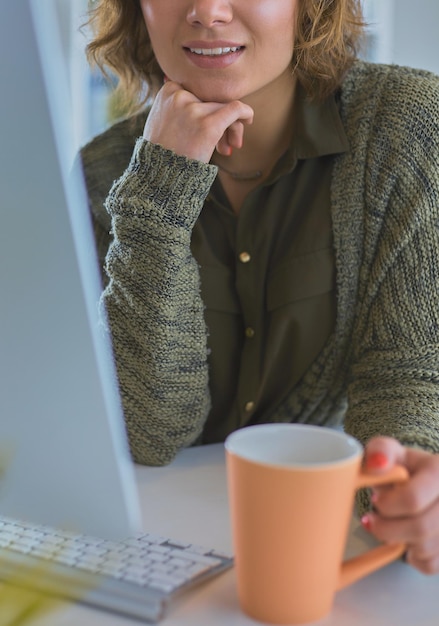 Attraktive lächelnde Frau, die am Schreibtisch sitzt und eine Tasse Kaffee hält, entspannt sich und schaut weg