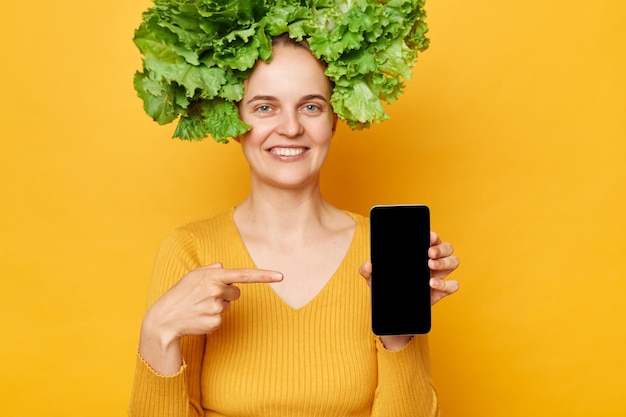 Attraktive lächelnde Frau in lässigem Hemd und Salatkranz steht isoliert vor gelbem Hintergrund und zeigt auf ihr Smartphone mit leerem Bildschirm für Werbung