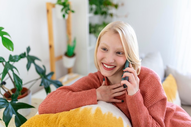 Attraktive lächelnde Frau mit Smartphone, während sie zu Hause auf dem Sofa sitzt. Kommunikations- und Gemütlichkeitskonzept.