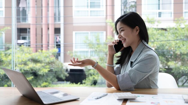 Attraktive lächelnde junge asiatische Geschäftsfrau, die sich im Büro entspannt und an einem Laptop arbeitet, der am Handy spricht