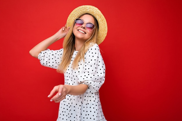 Attraktive lächelnde junge blonde Frau, die alltägliche stilvolle Kleidung und moderne Sonnenbrillen trägt, die auf einer bunten Hintergrundwand mit Blick auf die Kamera isoliert sind.