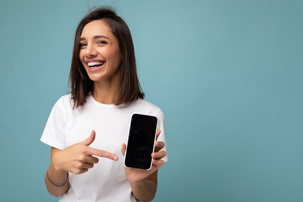 Attraktive lächelnde junge Brunet-Frau, die gut aussieht, trägt weißes T-Shirt, das isoliert auf blauem Hintergrund steht, mit Kopienraum, der Smartphone hält, das Telefon in der Hand mit leerer Bildschirmanzeige für Moc zeigt