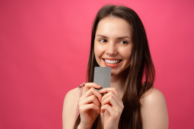 Attraktive lächelnde junge Frau, die schwarze Kreditkarte gegen rosa Studio hält