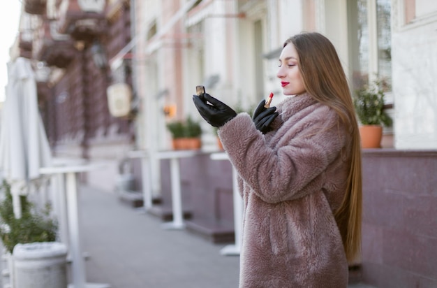 Attraktive Luxusfrau, die einen Pelzmantel trägt, malt ihre Lippen mit rotem Lippenstift in der Stadt Make-up