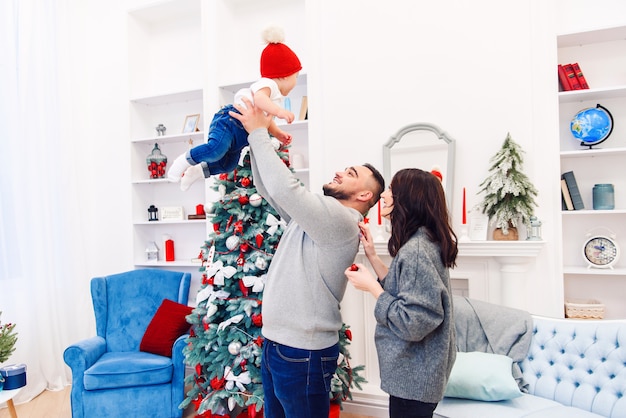 Attraktive Mama steht in der Nähe von hübschen wunderschönen Vater, die ihren entzückenden kleinen Sohn in der Luft in der Nähe von festlich geschmückten Weihnachtsbaum werfen.