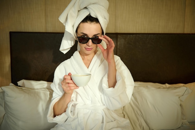 Attraktive modische, stilvolle hübsche Frau, die einen Bademantel trägt, mit ihrem Kopf in ein Badetuch gewickelt, auf dem Bett im Schlafzimmer sitzt, eine Tasse Kaffee hält und durch ihre Sonnenbrille in die Kamera schaut