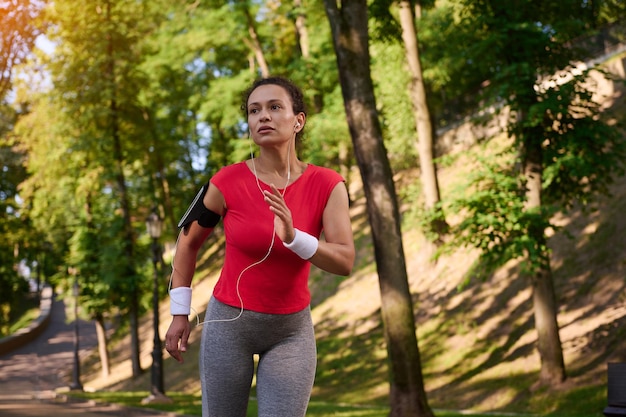 Attraktive, muskulöse, multiethnische Frau mit gesundem, schönem, ästhetischem Körper in enger Sportbekleidung mit Kopfhörern und Smartphone-Halterung, die auf dem Fußweg im Stadtpark Sport läuft