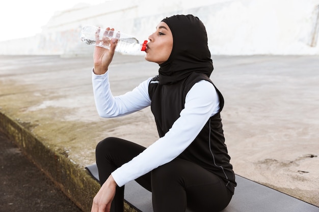 Attraktive muslimische Sportlerin, die im Freien Hijab trägt, auf einer Fitnessmatte sitzt und Wasser trinkt