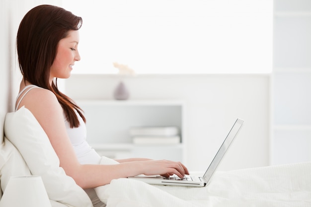 Attraktive redhaired Frau, die mit ihrem Laptop beim Sitzen auf ihrem Bett sich entspannt