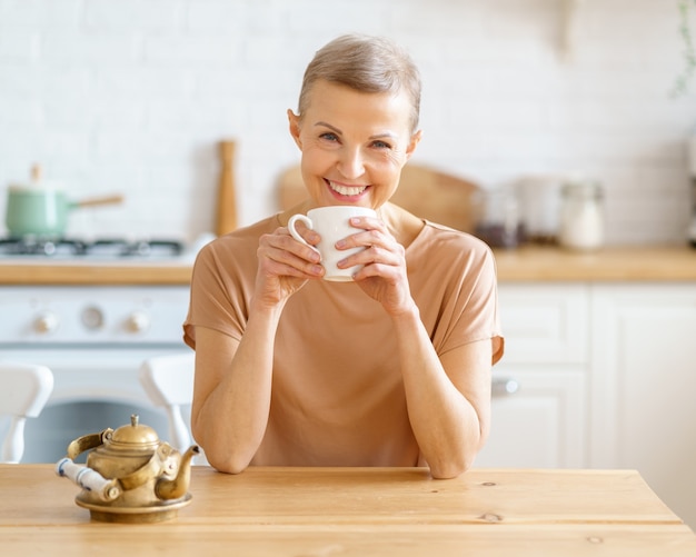 Attraktive reife Frau, die den Tag mit Kaffee beginnt, während sie am Holztisch in der Küche sitzt