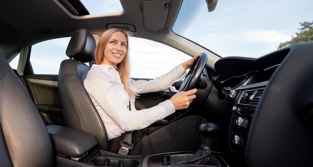 Attraktive reife Frau in formeller Kleidung, die sich mit dem Sicherheitsgurt anschnallt, während sie ein modernes Auto fährt Kaukasische Geschäftsfrau mit blonden Haaren, die ihren Weg zur Arbeit genießt