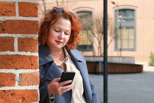 Attraktive rothaarige Frau in einem blauen Mantel hält ein Smartphone
