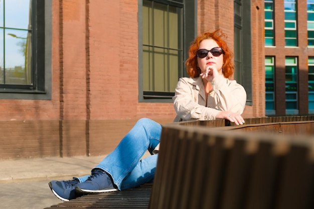 Attraktive rothaarige Frau mit Sonnenbrille sitzt auf einer Bank in einem Geschäftsviertel