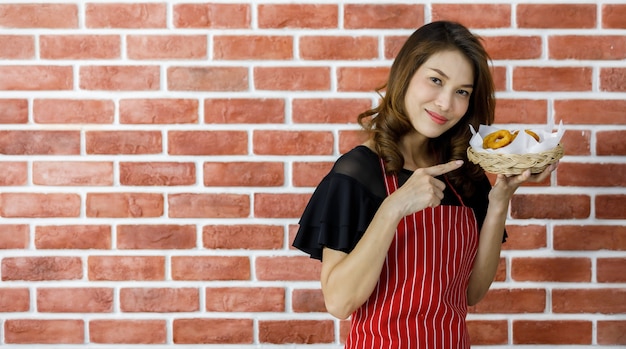Attraktive schöne junge asiatische Frau in rot gestreifter Schürze, die in der Nähe der Mauer steht und lächelnd überredet, köstliche Lieblingsspeisen zu essen, indem sie gebratenen Snack und sein Tablett in die Nähe des Mundes hebt