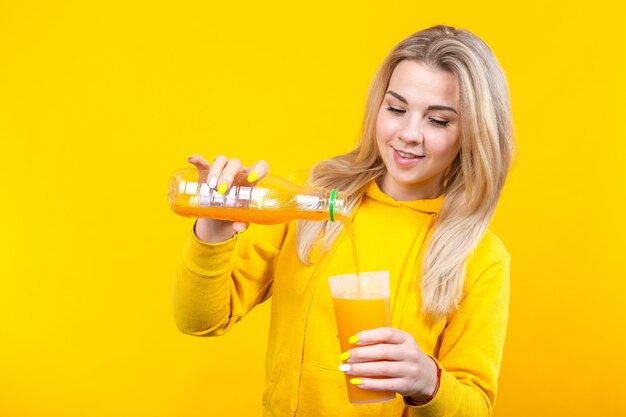 Attraktive schöne junge blonde Frau, die Orangensaft von Plastikflasche zu Glas gießt