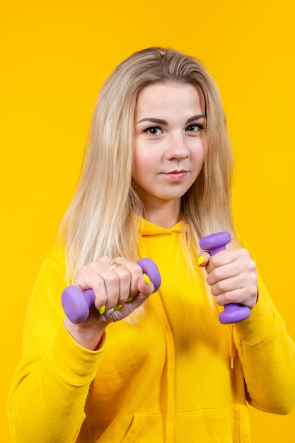 Attraktive schöne junge blonde Frau in lässiger gelber sportlicher Kleidung, die Übungen mit kleinen 0,5 kg lila Hanteln macht.