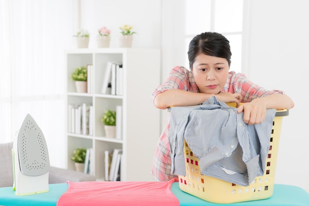 Foto attraktive schönheit hausfrau lehnt sich an kleiderkorb müde vom bügeln sortiert familienkleidung aus und benommen tagträumen steht im wohnzimmer.