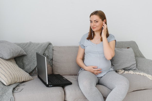 Attraktive schwangere Frau, die Bauch berührt und Laptop auf dem Bett benutzt