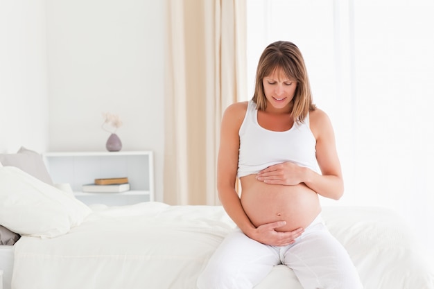 Attraktive schwangere Frau, die ihren Bauch beim Sitzen auf einem Bett streichelt