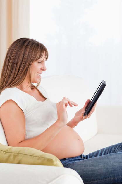 Attraktive schwangere Frau, die mit einer Computertablette beim Sitzen auf einem Sofa sich entspannt