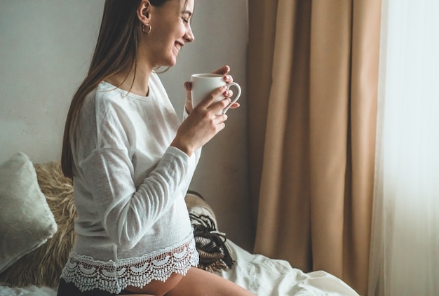 Attraktive schwangere frau trinkt tee auf dem bett. tee trinken, der durch ein fenster zu hause schaut. letzte monate der schwangerschaft.