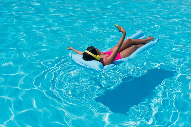 Attraktive schwarze Frau in Badebekleidung steht in der Sonne mit Handy in der Hand und Kopfhörern im tropischen Urlaub.