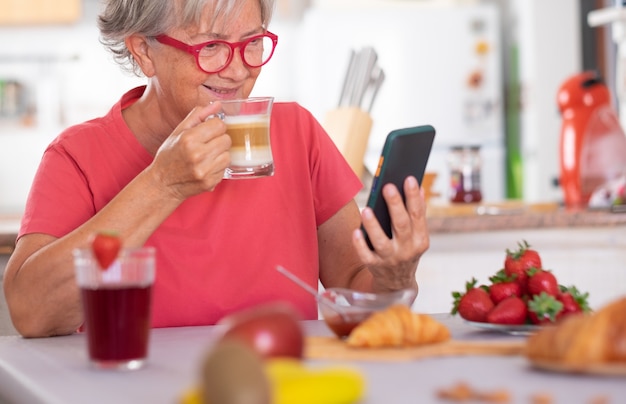 Attraktive Seniorin im Ruhestand, die zu Hause mit Croissant und Cappuccino frühstückt. Nachrichten auf dem Handy ansehen